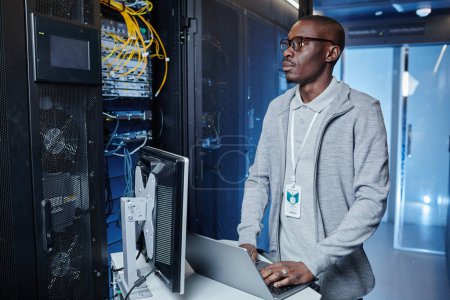 Téléchargez les photos : Portrait taille haute de l'homme noir adulte travaillant comme ingénieur informatique à l'aide d'un ordinateur portable tout en mettant en place un réseau Internet dans la salle des serveurs - en image libre de droit