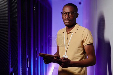 Téléchargez les photos : Portrait taille haute de l'homme afro-américain inspectant le réseau de données dans la salle des serveurs éclairée par la lumière au néon et regardant la caméra, l'espace de copie - en image libre de droit
