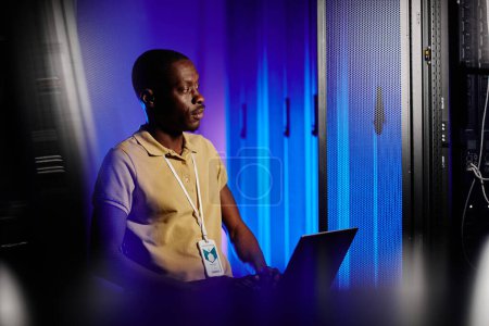 Téléchargez les photos : Portrait latéral d'un homme noir adulte utilisant un ordinateur portable dans une salle de serveurs sombre éclairée par une lumière au néon, espace de copie - en image libre de droit