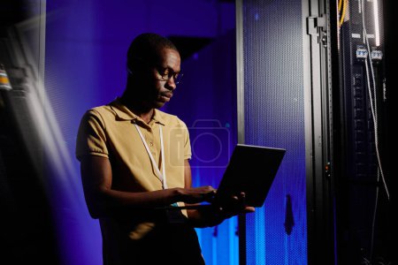 Téléchargez les photos : Portrait latéral d'un homme afro-américain adulte utilisant un ordinateur portable dans une salle de serveur sombre éclairée par une lumière au néon, espace de copie - en image libre de droit