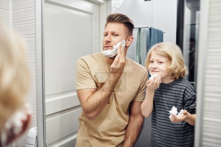Téléchargez les photos : Portrait de père et fils se rasant ensemble en regardant dans le miroir pendant la routine du matin dans la salle de bain - en image libre de droit