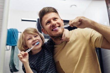 Retrato de bajo ángulo de padre e hijo juguetones cepillándose los dientes juntos por la mañana y mirándose en el espejo