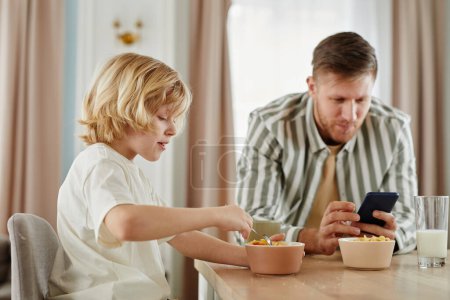 Photo for Side view portrait of father and son at breakfast in morning with dad using smartphone in background, copy space - Royalty Free Image