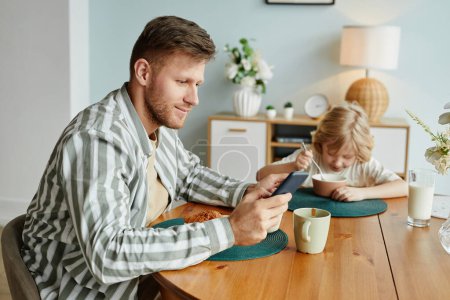 Téléchargez les photos : Portrait latéral de père utilisant un smartphone à table tout en prenant le petit déjeuner avec son fils - en image libre de droit