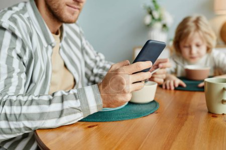 Téléchargez les photos : Gros plan du père à l'aide d'un smartphone à la table à manger avec un petit garçon en arrière-plan, espace de copie - en image libre de droit