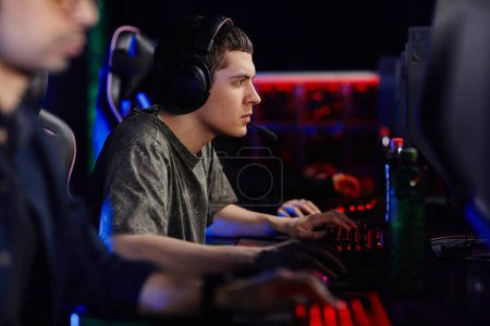 Photo for Side view portrait of focused young man playing video game with cyber sport team in dark - Royalty Free Image