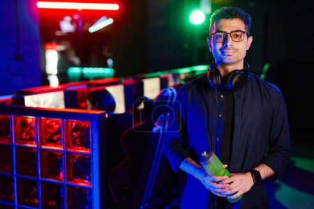 Photo for Waist up portrait of Middle Eastern young man wearing glasses in cyber sports club smiling at camera lit by neon light, copy space - Royalty Free Image