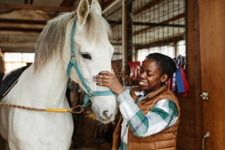 Foto de Retrato de una joven afroamericana acariciando un caballo blanco en establos y sonriendo felizmente - Imagen libre de derechos