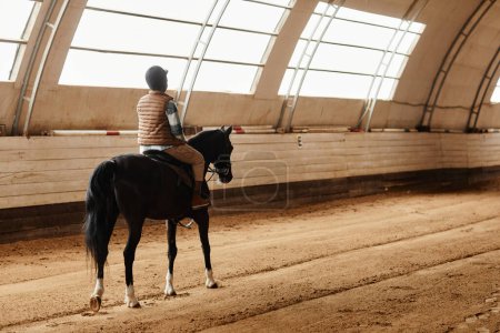 Foto de Amplia vista del ángulo en la mujer joven a caballo en la arena interior iluminada por la luz suave, espacio de copia - Imagen libre de derechos