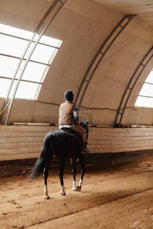 Foto de Vista posterior vertical a la mujer joven montar a caballo en la arena interior en el rancho de caballos o estadio de práctica - Imagen libre de derechos