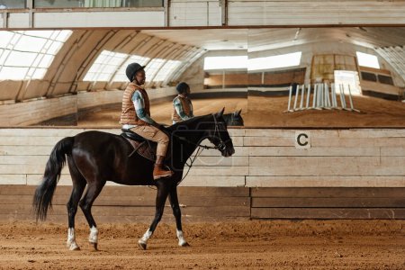 Foto de Vista lateral de longitud completa en la mujer joven montar a caballo en la arena interior o estadio de práctica con espejos, espacio para copiar - Imagen libre de derechos