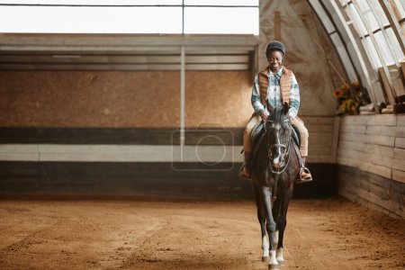 Foto de Retrato de larga duración de una joven sonriente montando a caballo en la arena interior en el rancho de caballos o el estadio de práctica, espacio para copiar - Imagen libre de derechos
