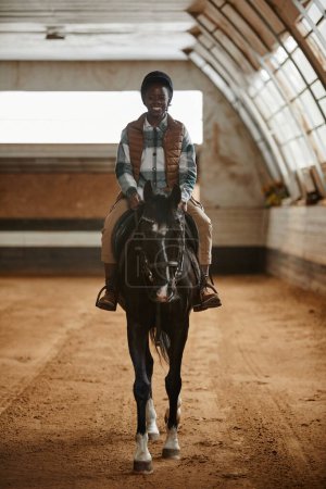 Photo for Vertical portrait of smiling young woman riding horse in indoor arena at horse ranch or practice stadium - Royalty Free Image