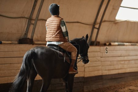 Téléchargez les photos : Vue arrière de la femme afro-américaine à cheval dans l'arène intérieure au ranch de chevaux ou au stade de pratique, espace de copie - en image libre de droit
