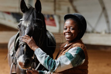 Foto de Retrato de la cintura hacia arriba de la mujer negra sonriente posando con el caballo en la arena de montar en interiores y mirando a la cámara, espacio de copia - Imagen libre de derechos