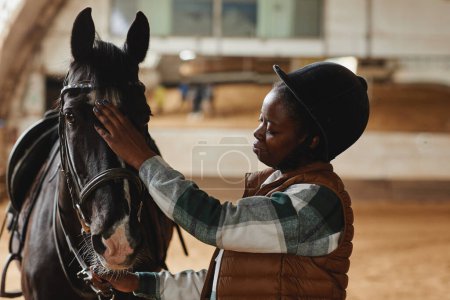 Photo for Side view portrait of young woman stroking horse while standing in indoor riding arena at practice - Royalty Free Image