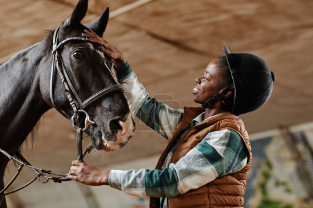 Foto de Retrato de vista lateral de una mujer afroamericana sonriente acariciando un caballo negro en la arena de la equitación interior en la práctica - Imagen libre de derechos