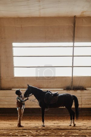 Foto de Retrato vertical de la mujer acariciando el caballo negro en la arena interior de la equitación durante la práctica, espacio de copia - Imagen libre de derechos