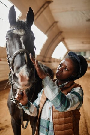 Foto de Retrato vertical de una joven mujer negra con caballo en la arena de la equitación interior después de la práctica de deportes ecuestres - Imagen libre de derechos