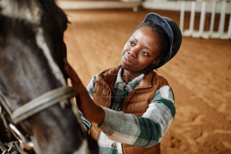 Téléchargez les photos : Portrait grand angle de jeune femme noire caressant le cheval après la pratique sportive dans l'arène d'équitation intérieure, espace de copie - en image libre de droit