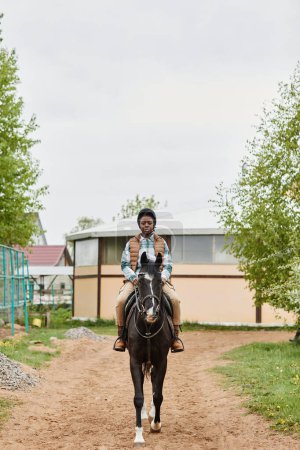 Téléchargez les photos : Portrait vertical pleine longueur de jeune femme noire chevauchant cheval vers la caméra dans un ranch extérieur - en image libre de droit