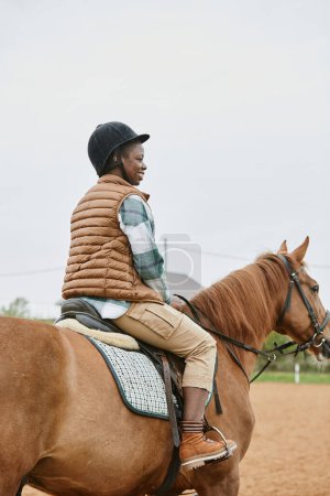 Photo for Vertical back view at young woman riding horse in ranch and wearing helmet - Royalty Free Image
