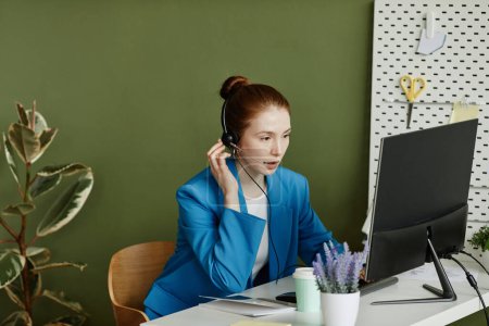 Téléchargez les photos : Jeune femme d'affaires dans un casque assis à table devant un écran d'ordinateur et parlant à ses collègues en ligne - en image libre de droit
