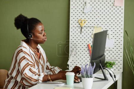 Photo for African young businesswoman in headphones sitting at her workplace with computer monitor and talking online with colleagues - Royalty Free Image