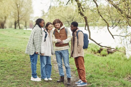 Téléchargez les photos : Groupe d'adolescents multiethniques utilisant une tablette numérique pour trouver le chemin lors de leur promenade dans le parc - en image libre de droit