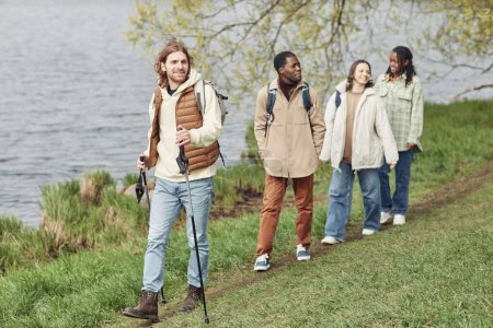 Téléchargez les photos : Jeune homme marchant avec bâton le long de la rive de la rivière avec ses amis derrière lui - en image libre de droit