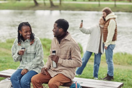 Téléchargez les photos : Jeune couple africain assis sur un banc et buvant du thé dans une fiole avec leurs amis faisant du selfie en arrière-plan au pique-nique en plein air - en image libre de droit
