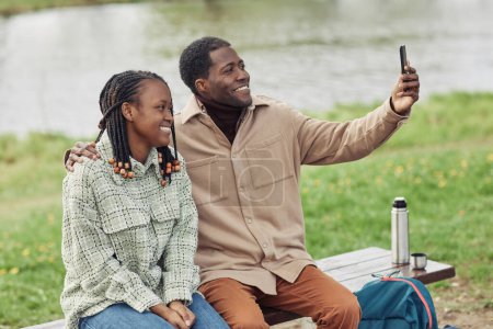 Téléchargez les photos : Heureux couple africain souriant à la caméra faisant selfie portrait sur téléphone portable tout en étant assis sur le banc dans le parc - en image libre de droit