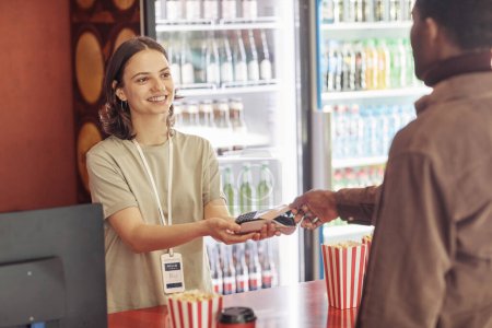 Photo for Young smiling seller holding terminal while customer paying for popcorn with credit card in the shop - Royalty Free Image