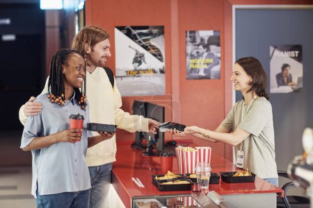 Téléchargez les photos : Jeune couple heureux achetant du pop-corn et du soda et payant par carte de crédit au cinéma - en image libre de droit