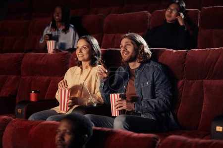 Téléchargez les photos : Jeune couple mangeant du pop-corn et regardant un film ensemble sur des fauteuils au cinéma - en image libre de droit