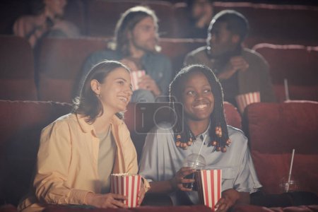 Téléchargez les photos : Deux femmes heureuses mangeant du pop-corn et regardant la comédie sur des chaises confortables dans l'auditorium - en image libre de droit