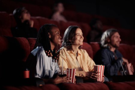 Photo for Two happy girls eating popcorn and laughing during watching comedy in the cinema - Royalty Free Image