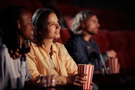 Téléchargez les photos : Jeune fille souriante manger du pop-corn tout en regardant un film avec son ami au cinéma - en image libre de droit