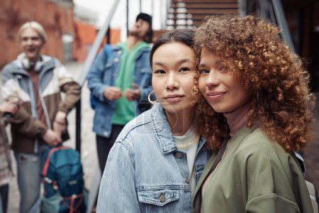 Téléchargez les photos : Deux jeunes femmes heureuses en tenues décontractées élégantes regardant la caméra tout en se tenant contre les adolescents qui bavardent par un escalier à l'extérieur - en image libre de droit
