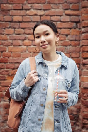 Photo for Young pretty woman in casualwear holding backpack on shoulder and soda in hand while standing against brick wall of building - Royalty Free Image
