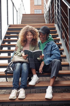 Téléchargez les photos : Couple adolescent souriant en tenue décontractée regardant à travers les notes de cours dans le copybook tout en étant assis sur un long escalier par un bâtiment moderne - en image libre de droit