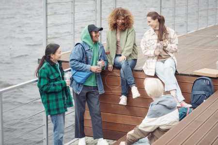 Photo for Five cheerful teenage friends in casualwear telling jokes or discussing funny things while having rest on wooden pier by waterside after college - Royalty Free Image