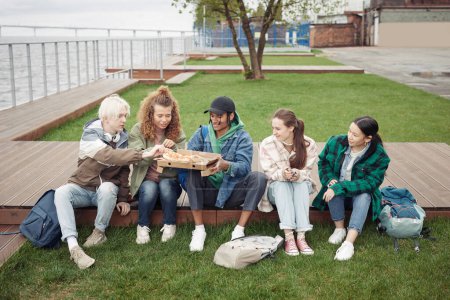 Foto de Grupo de amigos adolescentes interculturales sentados en el muelle de madera junto al césped verde y comiendo pizza mientras el tipo negro sostiene la caja cuadrada - Imagen libre de derechos
