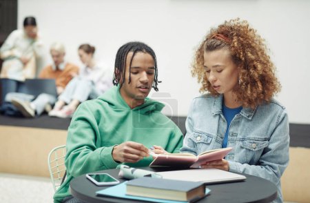 Foto de Adolescente y su novia discutiendo notas en copybook antes de la lección o seminario mientras están sentados a la mesa contra sus compañeros de clase - Imagen libre de derechos