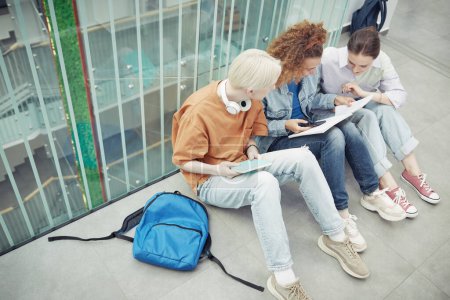 Photo for Three intercultural teenagers in casualwear looking through notes in copybook of girl with wavy hair while resting on the floor - Royalty Free Image