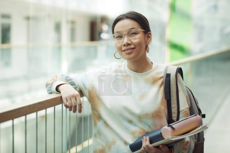 Foto de Sonriente adolescente con gafas y sudadera blanca sosteniendo suministros de educación mientras está de pie en el pasillo de la universidad en el descanso - Imagen libre de derechos