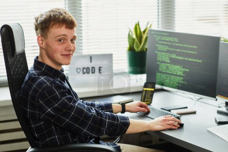 Foto de Retrato del joven programador mirando a la cámara mientras está sentado en su lugar de trabajo con un monitor de computadora y códigos de escritura - Imagen libre de derechos