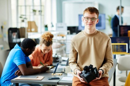 Téléchargez les photos : Portrait de jeune programmeur en lunettes tenant des lunettes de réalité virtuelle regardant la caméra tout en rencontrant ses collègues - en image libre de droit