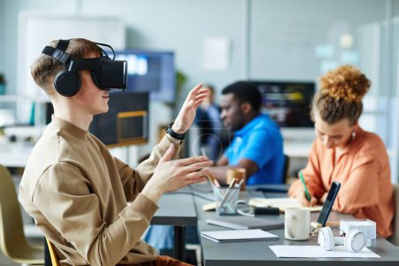 Photo for Young man in virtual reality glasses sitting at meeting with colleagues and testing new VR application - Royalty Free Image