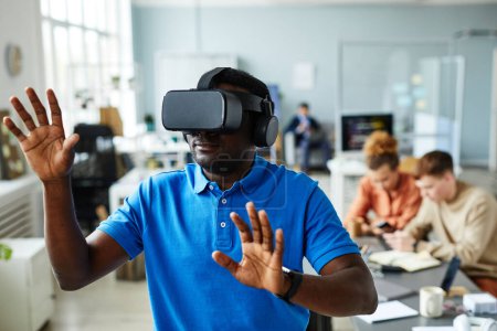 Photo for African young man in VR glasses gesturing while testing virtual reality program at office during meeting with colleagues - Royalty Free Image